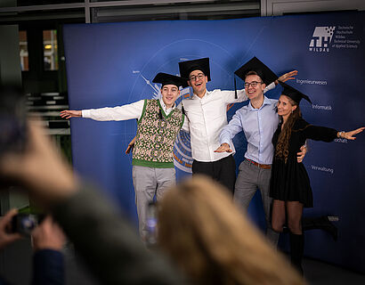 Feierliche Verabschiedung 2024 Absolvent*innen stehen für ein Foto sich umarmend nebeneinander vor einer blauen Wand und lächeln in die Kamera