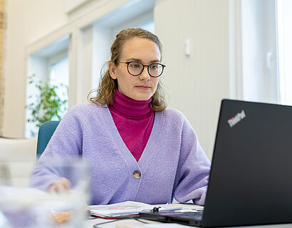 Aus Untersicht ist eine Frau zu sehen, die konzentriert an ihrem Laptop arbeitet. Im Vordergrund ist unscharf ein Glas Wasser zu erkennen.