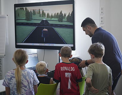 eine Gruppe von Kindern beobachtet ein weiteres Kind mit VR Brille beim virtuellen Autofahren