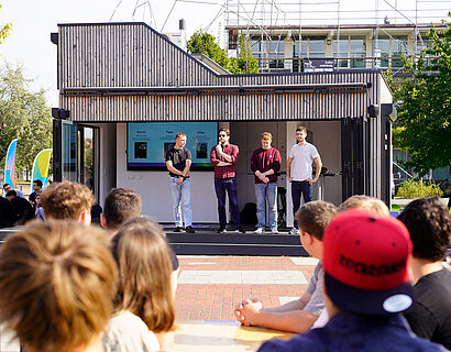 Vier Personen stehen vor der ScienceBox auf dem Campus der TH Wildau und begrüßen Studierende, welche im Vordergrund zu sehen sind