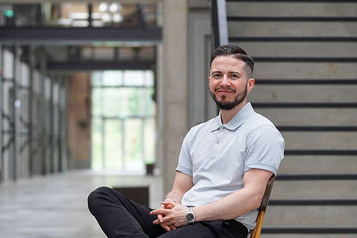 Ray Trautwein Person mit dunklen Haaren und Bart sitzt auf einem Stuhl in Halle 21 de TH Wildau und lächelt in die Kamera