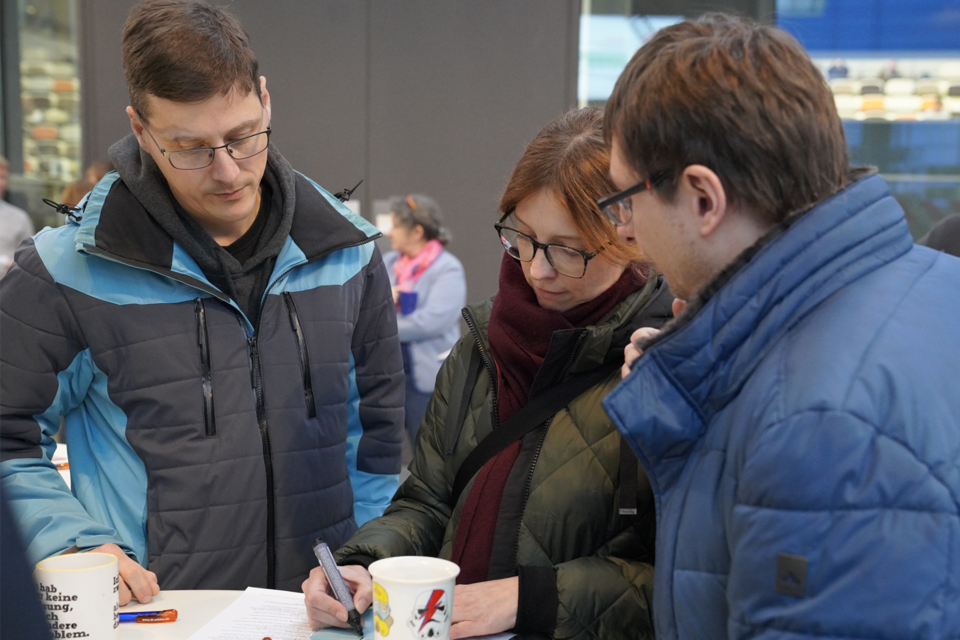 Drei Menschen stehen an einem Tisch und eine Person schreibt