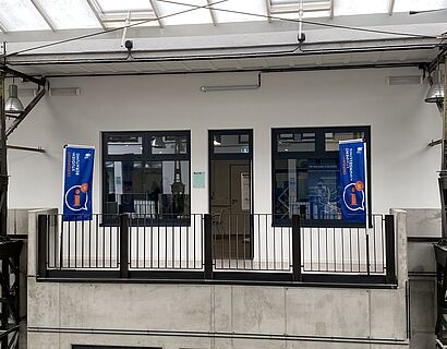 Außenansicht des Infopoints Studienberatung in Halle 21 auf dem Campus mit zwei Beachflags davor.