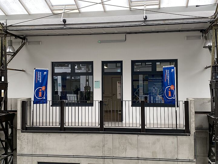 Außenansicht des Infopoints Studienberatung in Halle 21 auf dem Campus mit zwei Beachflags davor.