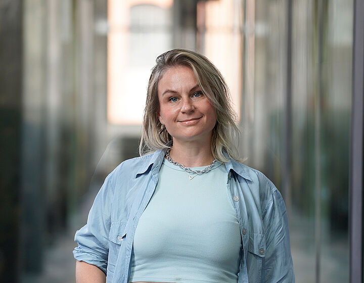 Iana Maksimova Portraitbild einer blonden Frau mit hellblauem Oberteil und blauem Hemd in einer Halle der TH Wildau