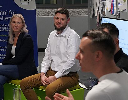 Im Vordergrund spricht ein Alumnus und gestikuliert. Im Hintergrund sind zwei weitere Alumni zu sehen, die ihm zuhören.