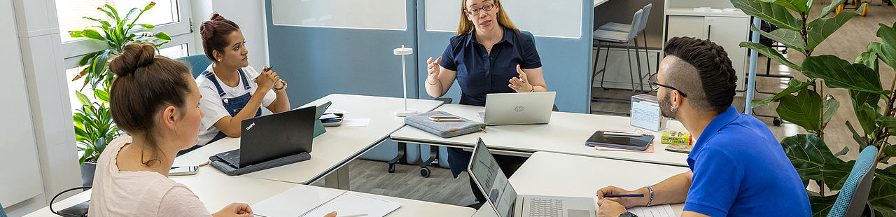 Eine Professorin und drei Studierende sitzen gemeinsam an einer Tischgruppe, die Professorin erklärt etwas.