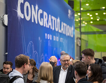 Absolvent*innen und Dozierende stehen vor einem blauen Banner mit der Aufschrift "Congratulations" im Foyer des Audimax