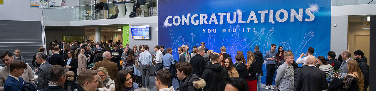 Absolvent*innen, Dozierende und Besucher*innen stehen im Foyer des Audimax vor einem blauen Banner mit der Aufschrift "Congratulations" und sind im Gespräch