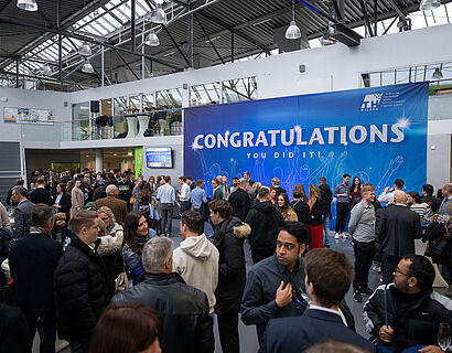 Absolvent*innen, Dozierende und Besucher*innen stehen im Foyer des Audimax vor einem blauen Banner mit der Aufschrift "Congratulations" und sind im Gespräch