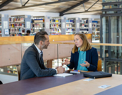 Eine Frau und ein Mann sitzen an einem Tisch in der Bibliothek und unterhalten sich.