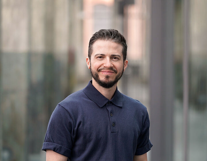 Ray Trautwein Person mit dunklem Shirt, schwarzen Haaren und Bart steht für ein Portraitbild in einer alten Industriehalle mit Betonpfeilern im Hintergrund.