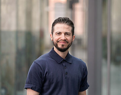 Ray Trautwein Person mit dunklem Shirt, schwarzen Haaren und Bart steht für ein Portraitbild in einer alten Industriehalle mit Betonpfeilern im Hintergrund.
